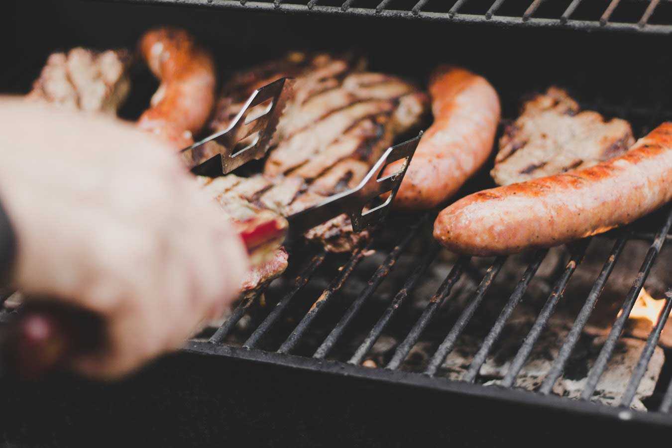Hand with Tongs Turning Steaks and Sausages on Barbecue