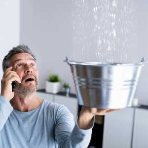 Man on the phone holding a silver bucket under a dripping leak