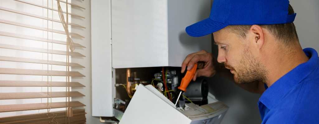a male plumber in a blue cap using a screwdriver inside an open boiler